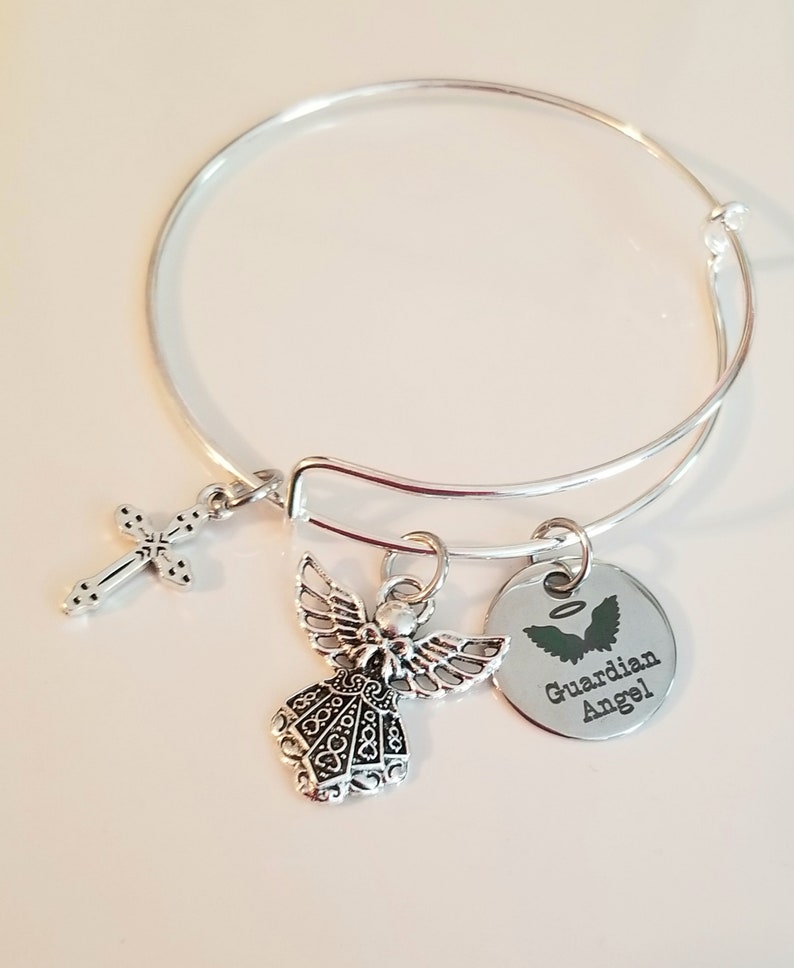 May include: Silver bangle bracelet with a charm of an angel, a cross, and a round charm with the text "Guardian Angel".