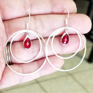 May include: Silver hoop earrings with a double-circle design. Each earring features a teardrop-shaped, faceted, red gemstone pendant. The earrings are suspended from a simple hook.