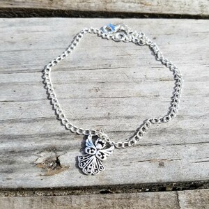 Op de afbeelding: Een zilverkleurige kettingarmband met een decoratieve engelhanger. De engelhanger heeft gedetailleerde vleugel- en lichaamsontwerpen. De armband wordt gepresenteerd op een verweerd houten oppervlak.