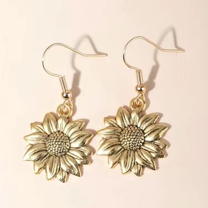 May include: A pair of gold-colored sunflower earrings. Each earring features a detailed sunflower pendant with a textured center and petals. The earrings have a simple hook design for easy wear. The background is a soft, neutral color.