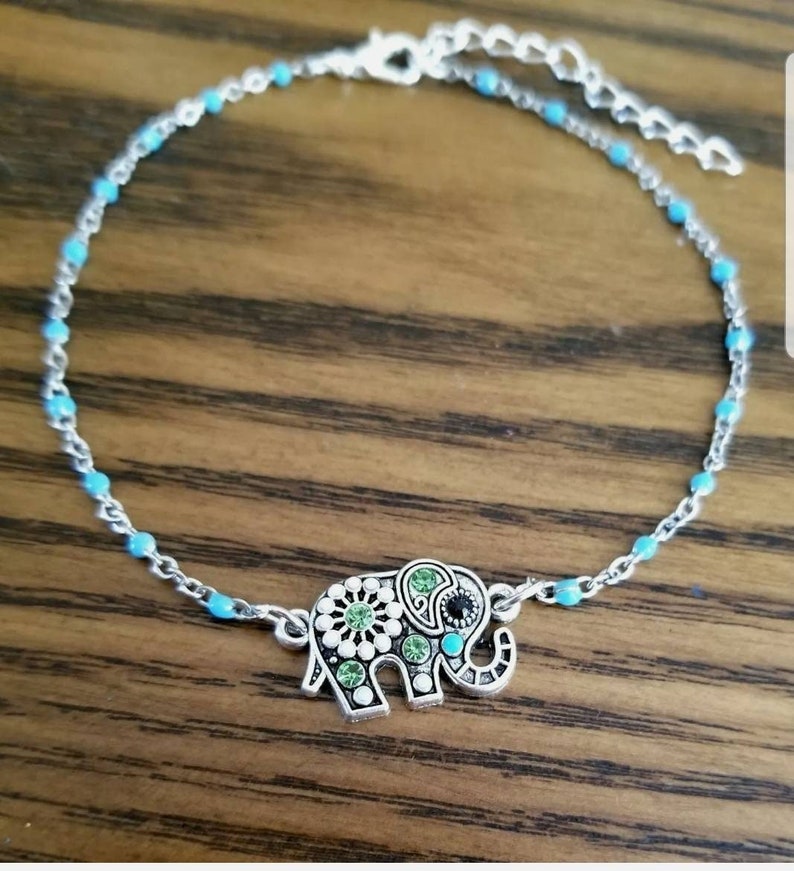 Op de afbeelding: Een zilveren enkelbandje met een turquoise blauwe ketting en een zilveren olifant bedel met groene en witte strass steentjes.
