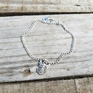 Op de afbeelding: Een zilverkleurige kettingarmband met een decoratieve engelbedel. De bedel heeft een engel met gedetailleerde vleugels en een halo. De armband ligt op een verweerd houten oppervlak.