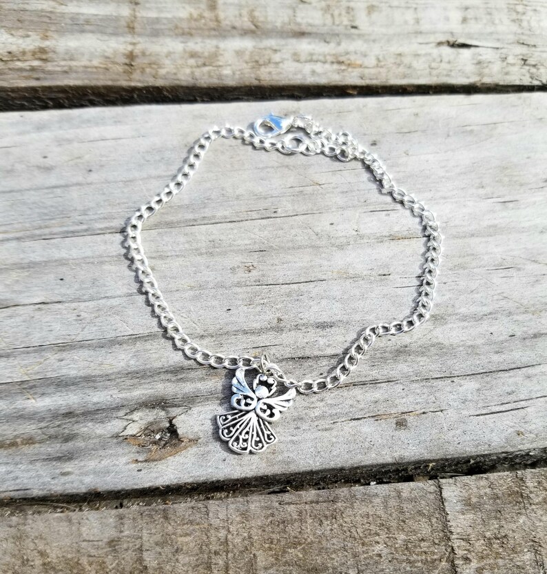 Op de afbeelding: Een zilverkleurige kettingarmband met een decoratieve engelbedel. De bedel heeft een engel met gedetailleerde vleugels en een halo. De armband ligt op een verweerd houten oppervlak.