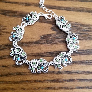 Op de afbeelding: Zilveren armband met zeven olifantenbedels. Elke olifantenbedel is versierd met groene, witte en blauwe strasssteentjes.