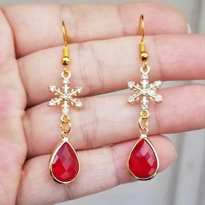 Könnte beinhalten: Goldfarbene Schneeflocken-Ohrringe mit roten tropfenförmigen Anhängern. Die Ohrringe haben ein Schneeflocken-Design mit klaren Kristallen und einem roten, facettierten Tropfen. Die Ohrringe hängen an einem goldfarbenen Haken.