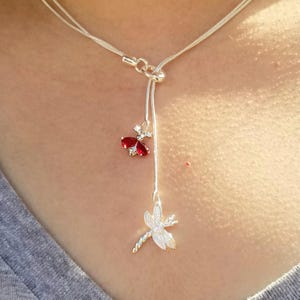 May include: A silver-toned lariat necklace featuring a delicate snake chain. The necklace includes two dangling charms: a small ladybug with red wings and a detailed dragonfly pendant with clear stone accents, creating a layered vertical design.