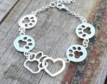 May include: Silver-toned bracelet featuring paw prints and a heart charm. The bracelet has a chain with a lobster clasp closure. The paw prints are in circular settings, and the heart charm is open. The bracelet is displayed on a wooden surface.