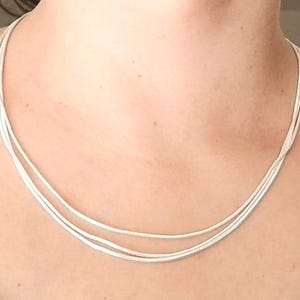 May include: A silver necklace with three strands, draped around a person's neck. The necklace appears to be made of thin, flexible metal strands, creating a delicate and elegant look. The necklace is a simple, yet stylish accessory.