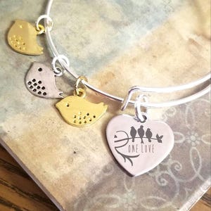 May include: Silver bangle bracelet with three bird charms and a heart charm with the words "One Love" and five birds on a branch.