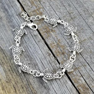 Könnte beinhalten: Silberfarbenes Armband mit einer Reihe detaillierter Schildkrötenanhänger. Das Armband hat einen Karabinerverschluss und eine verstellbare Kette. Die Schildkröten sind gleichmäßig verteilt und durch kleine, kreisförmige Glieder verbunden. Das Armband wird auf einer verwitterten Holzoberfläche präsentiert.
