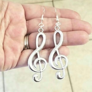 May include: A pair of silver-coloured treble clef earrings. The earrings have a hook at the top and a treble clef design. The earrings are made of a shiny metal and are displayed against a neutral background.