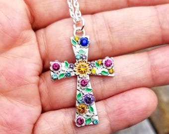 May include: Silver-tone cross pendant necklace with colorful floral and gemstone accents. The cross features a detailed design with pink, blue, yellow, and purple flowers and gemstones. The necklace is held in a hand.