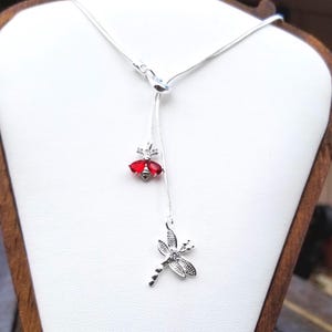 May include: A silver-toned lariat necklace featuring two dangling charms. One charm is a bee with red teardrop-shaped wings, and the other is a dragonfly with textured wings and a small clear stone accent. The chain is a thin, flexible snake style.