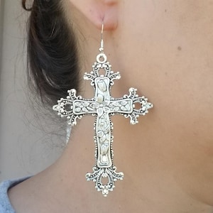 May include: Silver-toned cross earrings with intricate floral and scrollwork details. The earrings are large and dangle from a simple hook. The cross design is ornate and detailed, with a vintage or gothic aesthetic.
