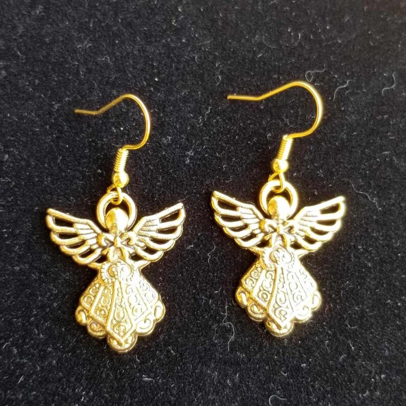 Puede incluir: Un par de pendientes dorados con forma de &aacute;ngel. Cada pendiente presenta un colgante de &aacute;ngel detallado con alas extendidas y un vestido fluido. Los pendientes tienen un cierre de gancho simple y se muestran sobre un fondo oscuro.