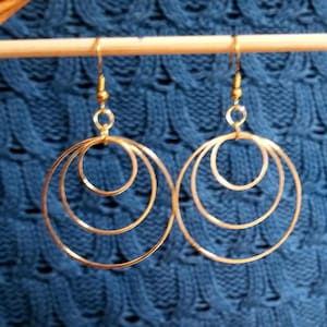 May include: A pair of gold-colored hoop earrings. Each earring features three nested circular hoops of decreasing size, suspended from a small hook. The earrings are displayed against a textured blue background.