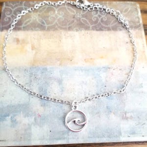 May include: A silver-colored anklet featuring a delicate chain and a circular pendant. The pendant showcases a wave design within a circle. The anklet is displayed on a decorative surface with a mix of blue, beige, and white tones.