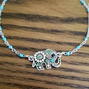 Op de afbeelding: Een zilveren enkelketting met een turquoise blauwe kralenontwerp en een zilveren olifantbedel met groene en zwarte strass stenen.