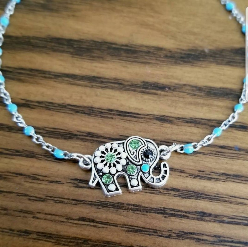 Op de afbeelding: Een zilveren enkelketting met een turquoise blauwe kralenontwerp en een zilveren olifantbedel met groene en zwarte strass stenen.