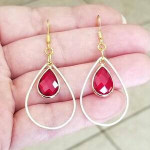 May include: A pair of gold-colored drop earrings. Each earring features a teardrop-shaped, faceted, red gemstone suspended within a gold-tone frame. The earrings are held up by a gold-colored hook.