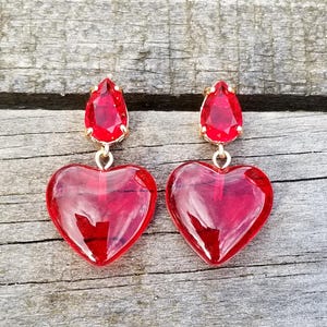 May include: A pair of heart-shaped red earrings with a gold-colored top. The earrings have a teardrop-shaped red gem at the top and a large, transparent heart pendant. The earrings are displayed on a wooden surface.