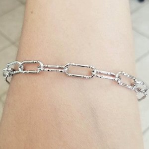 May include: A silver-colored chain bracelet with an elongated oval link design. The bracelet is worn on a person's arm, showcasing the intricate details of the links. The links have a textured pattern.