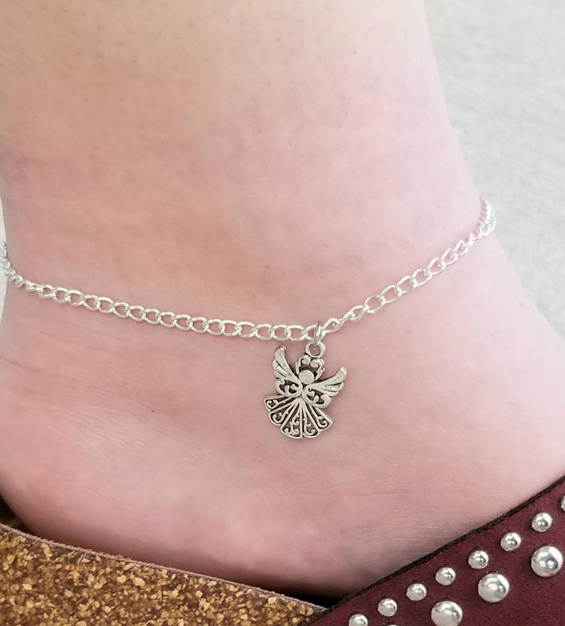Op de afbeelding: Een zilverkleurige enkelband met een kettingontwerp en een engelbedel. De engelbedel heeft gedetailleerde vleugels en een decoratieve rok. De enkelband wordt gedragen op een enkel met een bruine en rode sandaal.