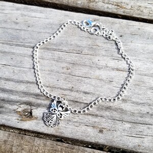 Op de afbeelding: Een zilverkleurige kettingarmband met een decoratieve engelbedel. De bedel heeft een open ontwerp. De armband wordt getoond op een verweerd houten oppervlak.