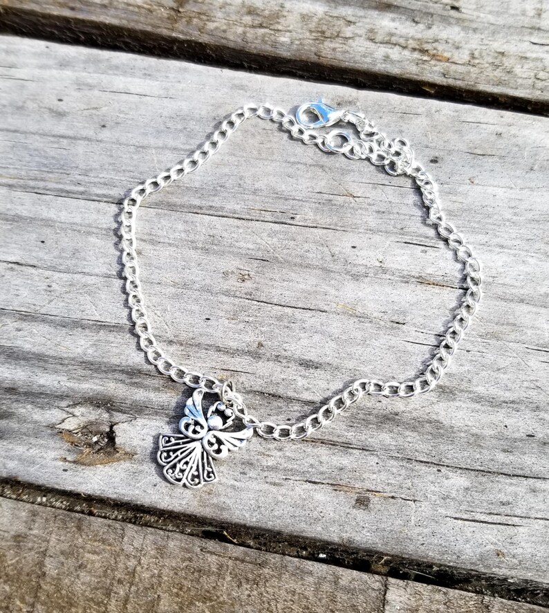 Op de afbeelding: Een zilverkleurige kettingarmband met een decoratieve engelbedel. De bedel heeft een open ontwerp. De armband wordt getoond op een verweerd houten oppervlak.