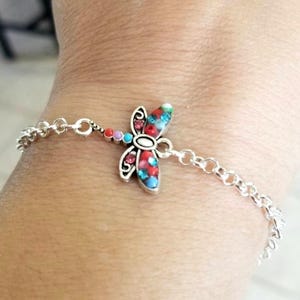 Op de afbeelding: Zilverkleurige armband met een libellenbedel. De libel heeft rode, blauwe en turquoise details. De armband heeft een fijn schakelontwerp en de libellenbedel is het middelpunt.