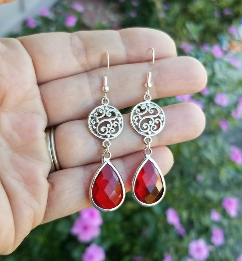 以下が含まれることがあります： 赤い雫型のクリスタルが付いたシルバーのフィリグリーイヤリング。イヤリングは繊細で複雑なデザインです。