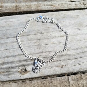 Op de afbeelding: Een zilverkleurige kettingarmband met een decoratieve engelbedel. De bedel toont een engel met gedetailleerde vleugels en een halo. De armband ligt op een verweerd houten oppervlak.