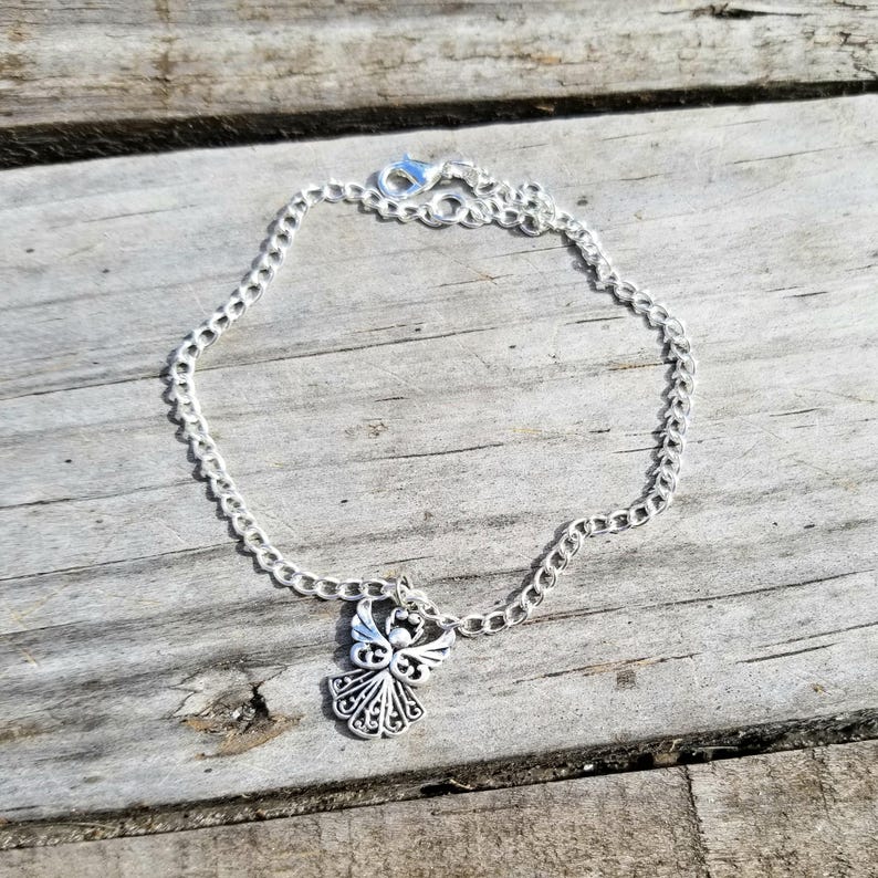 Op de afbeelding: Een zilverkleurige kettingarmband met een decoratieve engelbedel. De bedel toont een engel met gedetailleerde vleugels en een halo. De armband ligt op een verweerd houten oppervlak.