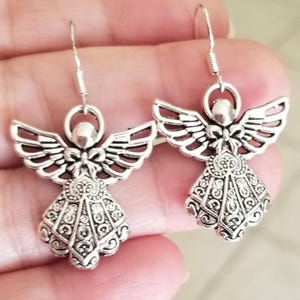 Puede incluir: Un par de pendientes plateados con forma de &aacute;ngel. Cada pendiente presenta un dise&ntilde;o de &aacute;ngel con alas detalladas, un halo y un vestido decorativo. Los pendientes tienen un cierre de gancho para orejas perforadas.
