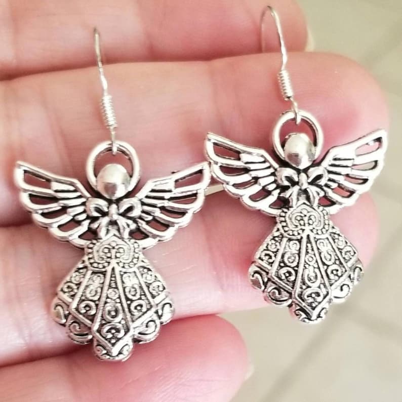 Puede incluir: Un par de pendientes plateados con forma de &aacute;ngel. Cada pendiente presenta un dise&ntilde;o de &aacute;ngel con alas detalladas, un halo y un vestido decorativo. Los pendientes tienen un cierre de gancho para orejas perforadas.