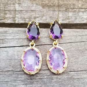May include: A pair of gold-toned drop earrings featuring a faceted pear-shaped purple gemstone at the top and a larger faceted oval-shaped light purple gemstone at the bottom, both set in a decorative gold-toned metal frame with a textured edge.