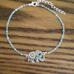 Op de afbeelding: Een zilveren enkelbandje met een turquoise blauwe kralenketting en een olifant bedel. De olifant bedel is versierd met groene en witte strass steentjes.