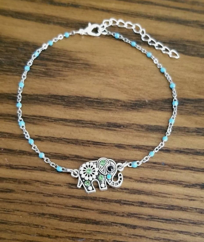 Op de afbeelding: Een zilveren enkelbandje met een turquoise blauwe kralenketting en een olifant bedel. De olifant bedel is versierd met groene en witte strass steentjes.