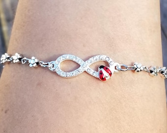 Puede incluir: Pulsera plateada con un símbolo de infinito incrustado con cristales transparentes y un pequeño dije de mariquita roja y negra. La pulsera también tiene pequeños detalles florales.