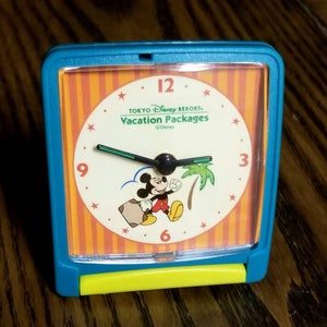 May include: A teal blue and yellow alarm clock with a Mickey Mouse design. The clock face has orange and white stripes and features Mickey Mouse carrying a suitcase and a palm tree. The clock face also says "Tokyo Disney Resort Vacation Packages" and "©Disney".
