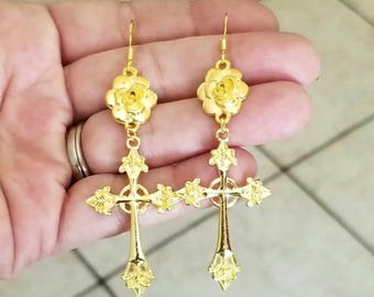 Puede incluir: Pendientes colgantes de tono dorado con un diseño de cruz y rosa.