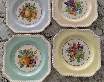 Puede incluir: Un juego de cuatro platos cuadrados de cerámica con esquinas redondeadas y un borde con textura de cuerda. Cada plato presenta un color pastel diferente—azul claro, crema, verde menta y amarillo pálido—y una ilustración central de frutas.