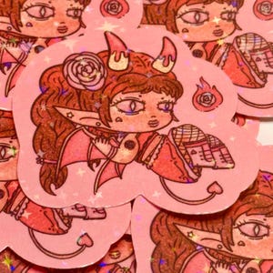 May include: A collection of pink stickers featuring a cartoon devil girl with brown hair, horns, and a tail. The stickers have a holographic effect and include a small flame and rose design.