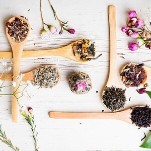 May include: Assortment of loose leaf teas displayed in wooden spoons on a white wooden background. The teas include green tea, black tea, herbal tea, and flower tea.