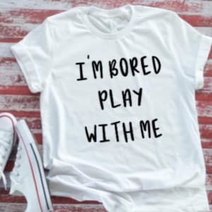 May include: White t-shirt with the text "I'M BORED PLAY WITH ME" in black. The shirt is paired with white sneakers with red and blue accents, set against a red and white striped wooden surface.