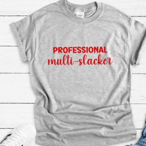 May include: A heather gray t-shirt with the words "PROFESSIONAL multi-slacker" printed in red. The shirt has a crew neck and short sleeves, with one sleeve rolled up. The shirt is laying on a white wooden surface.