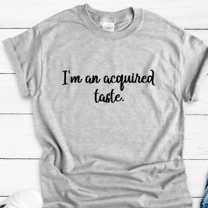 May include: A light gray t-shirt with the phrase "I'm an acquired taste" printed in black script across the front. The shirt has short sleeves and a crew neck.