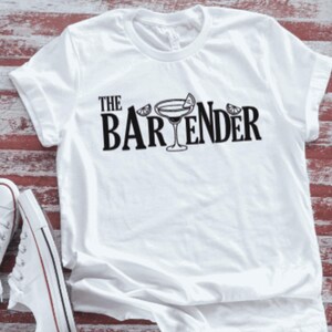 May include: White t-shirt with the text "THE BARTENDER" in black, featuring a cocktail glass illustration. The shirt has rolled-up sleeves and is paired with white sneakers. The background has a red and white striped pattern.
