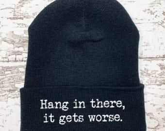 May include: Black beanie with the embroidered phrase "Hang in there, it gets worse." The hat is made of a soft, knitted material. The text is in a white, sans-serif font. The beanie is a classic, casual accessory.