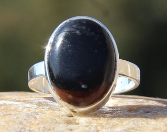 May include: A silver ring featuring a large, oval-shaped black gemstone. The ring's band is a polished silver, and the gemstone has a smooth, reflective surface. The ring is set against a neutral, textured background.